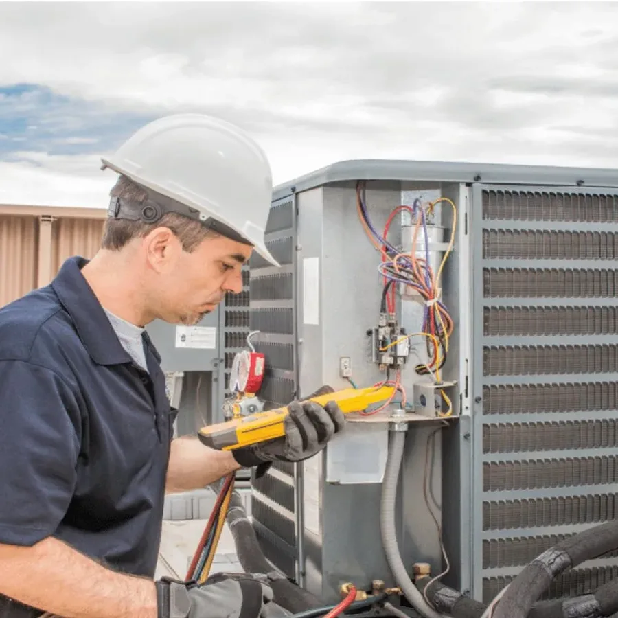 HVAC technician in a hard hat testing an outdoor air conditioner with a multimeter